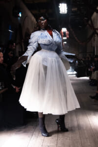 Model wears a blue and white striped blouse with a cinched corseted waist and a voluminous white tulle skirt on a dim runway at a fashion show.