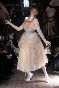 Models on a runway wearing a voluminous white satin gown with clear plastic overlay, white gloves, braided hair, and carrying a small white handbag.