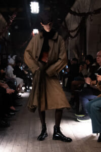 Model walks a runway in a tan trench coat, black turtleneck, and a burgundy waist belt, wearing black lace-up boots as spectators watch in dim lighting.