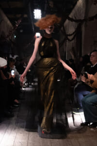 Model walks a runway in a metallic gold gown with a black sheer overlay; dramatic orange hair and spotlight overhead, audience on both sides filming.