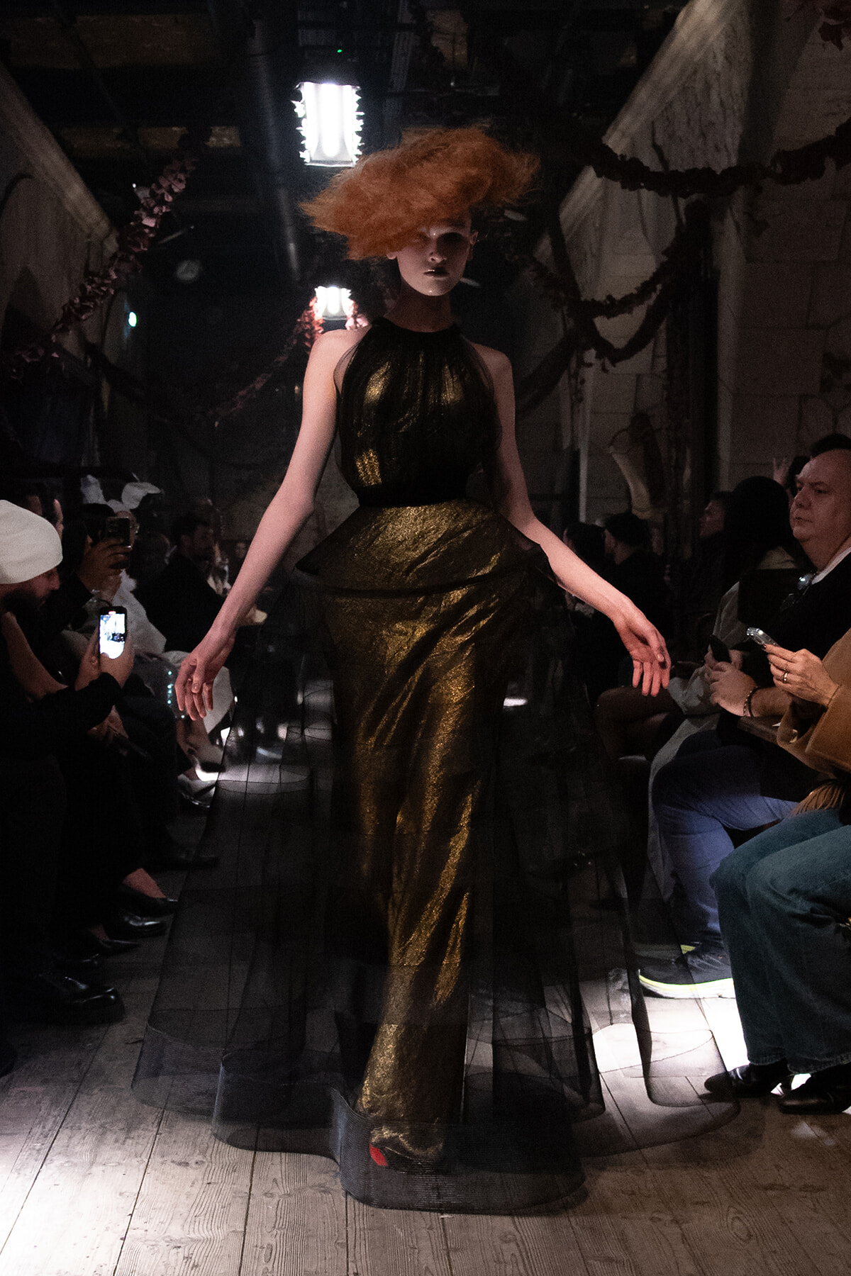 Model walks a runway in a metallic gold gown with a black sheer overlay; dramatic orange hair and spotlight overhead, audience on both sides filming.