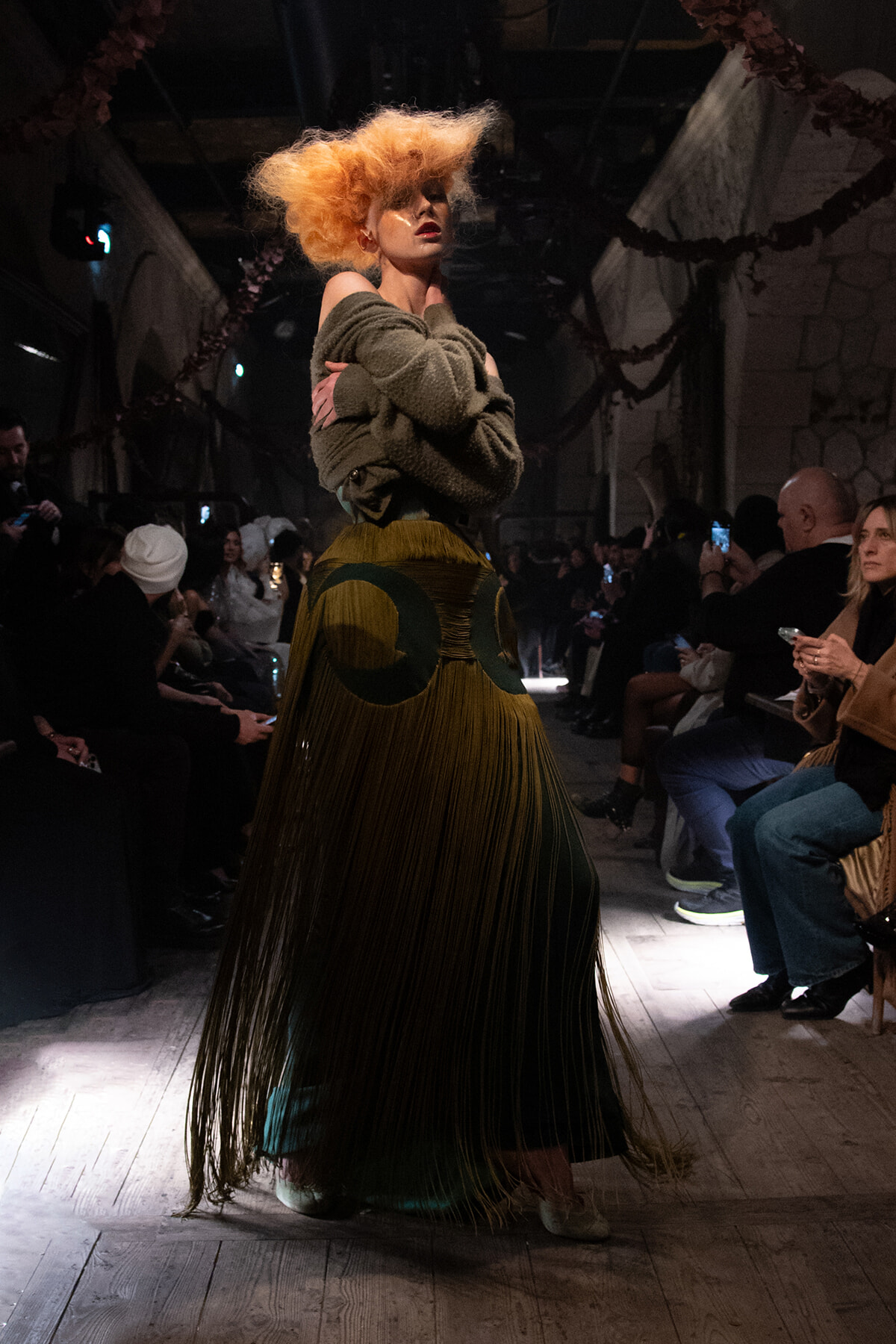 Model struts a runway wearing an oversized olive knit sweater and a long, fringed yellow-green skirt, with voluminous orange hair under dim venue lighting.