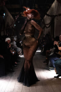 Model on a runway wearing a sheer black lace gown with a long train and dramatic red hair, surrounded by seated fashion show attendees.