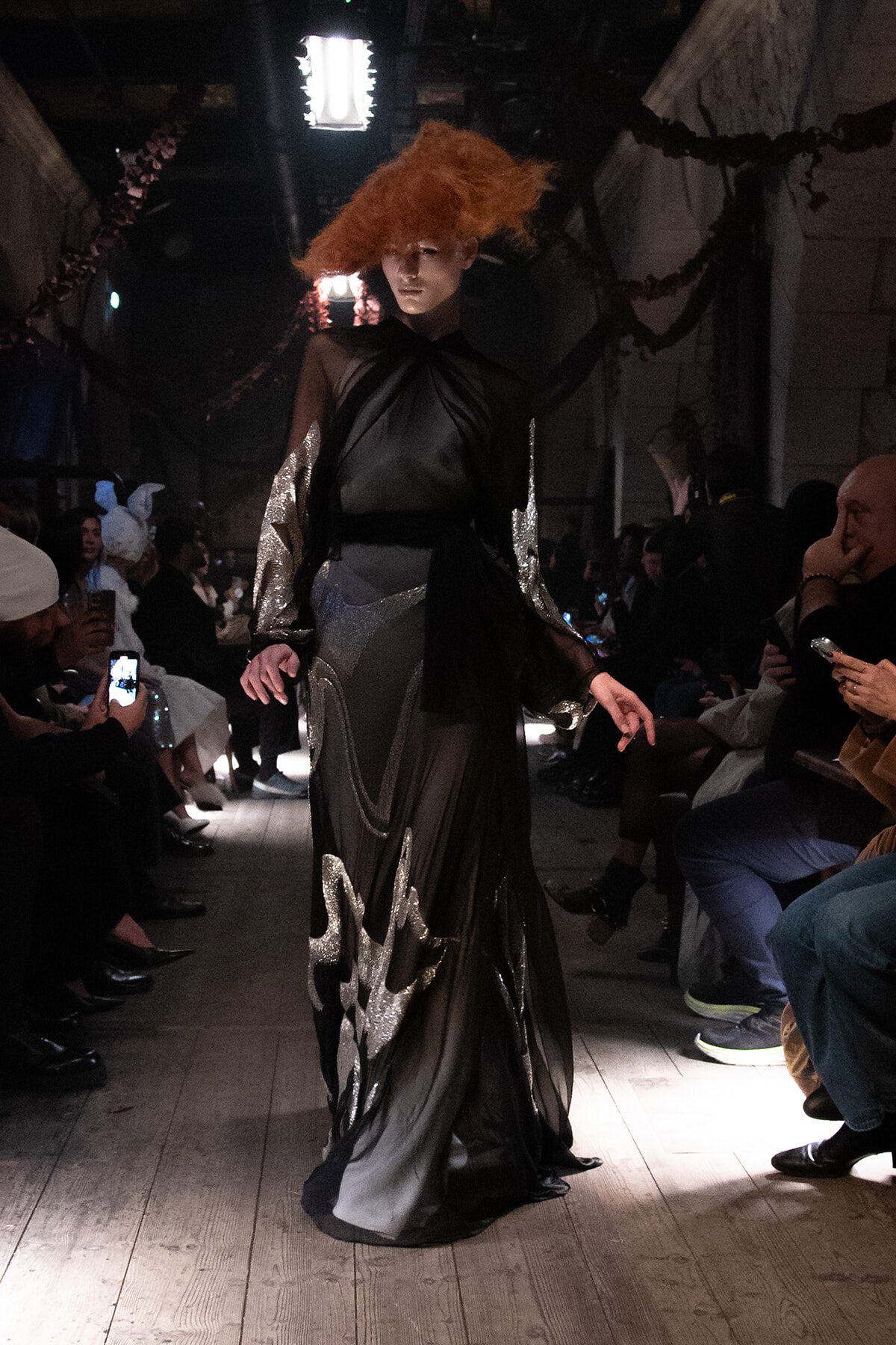 Model walks the runway in a black sheer gown with silver metallic accents as the audience watches under dim lighting on both sides.