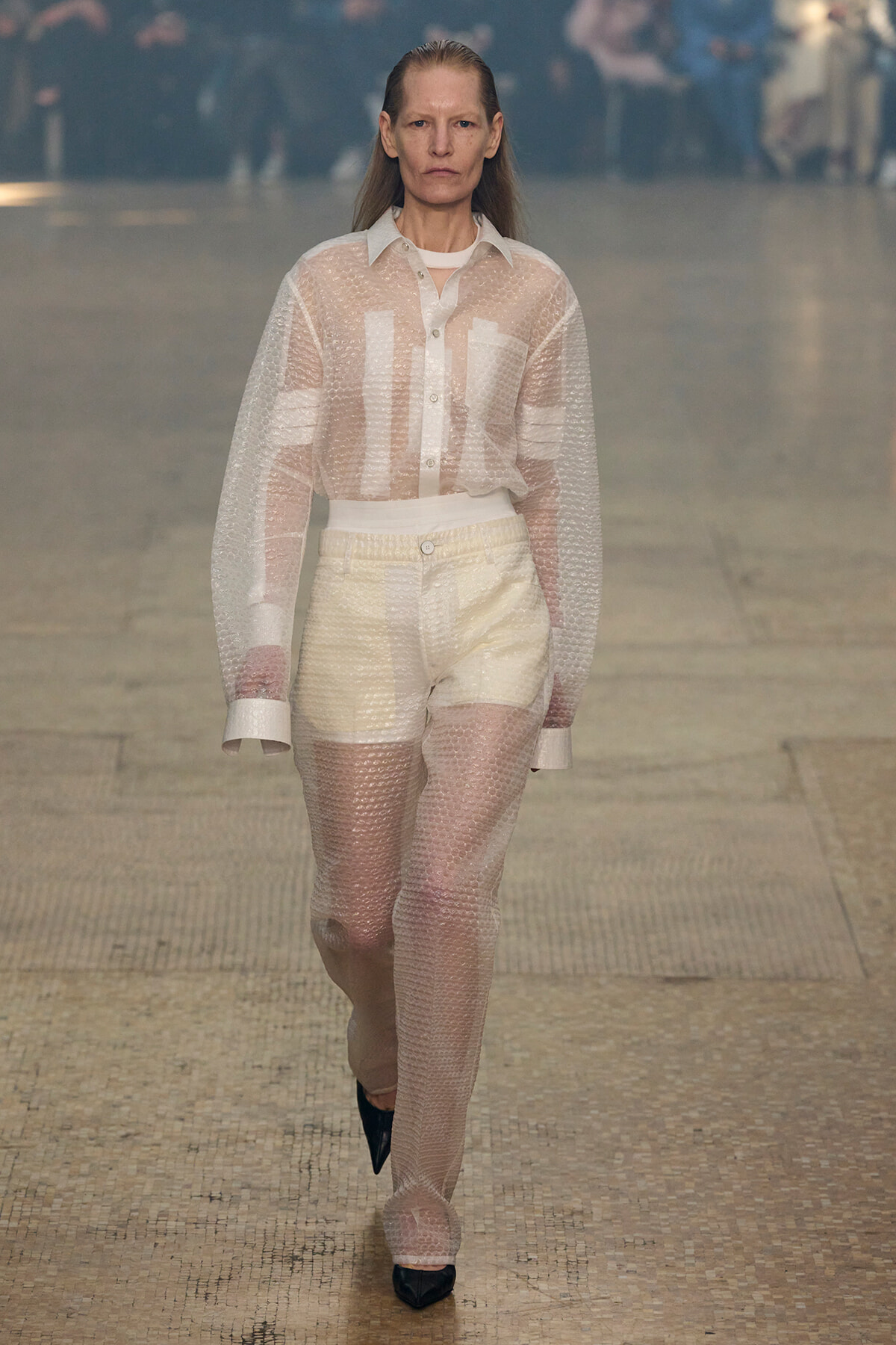 Model walking a runway in a sheer bubble-wrap outfit: white button-down shirt and high-waisted pants, transparent texture visible