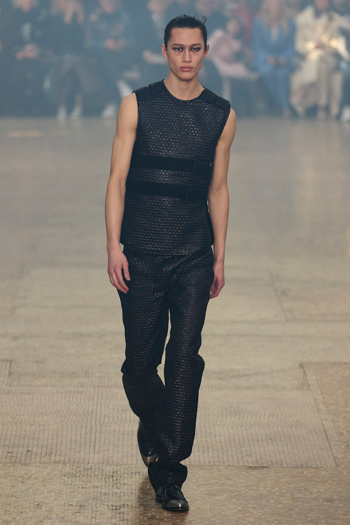 Model walks a runway in a sleeveless black textured outfit with a belt and matching pants, focused expression, audience blurred in the background.