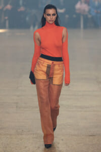 Model walking a runway in a bright orange turtleneck with shoulder cutouts and orange patchwork pants with a black belt? but no question marks.