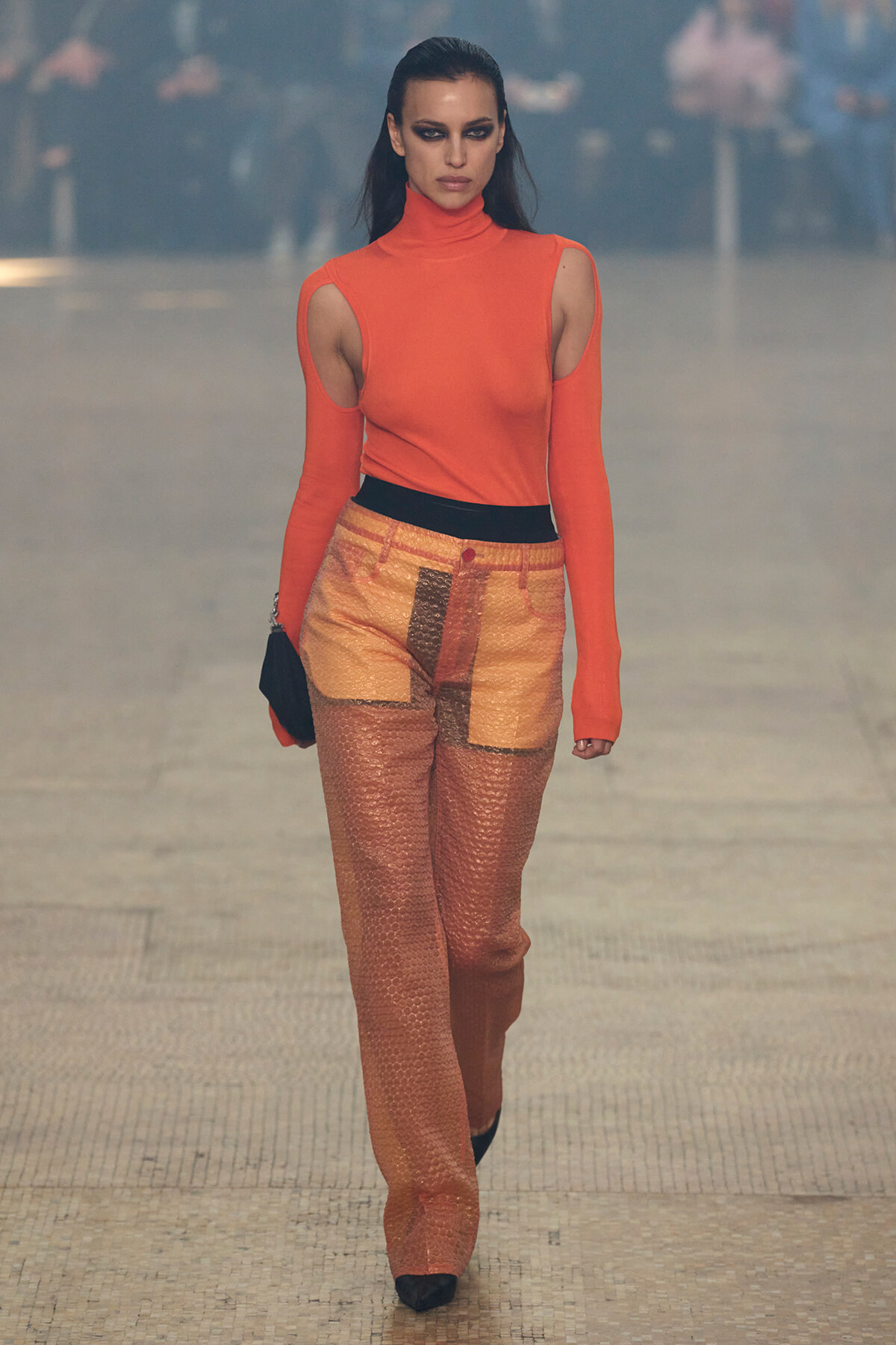 Model walking a runway in a bright orange turtleneck with shoulder cutouts and orange patchwork pants with a black belt? but no question marks.