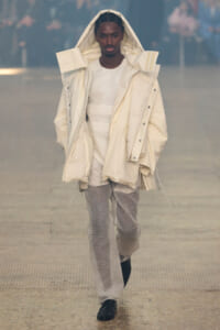 Model on a runway wearing an oversized cream hooded puffer coat over a white textured outfit.