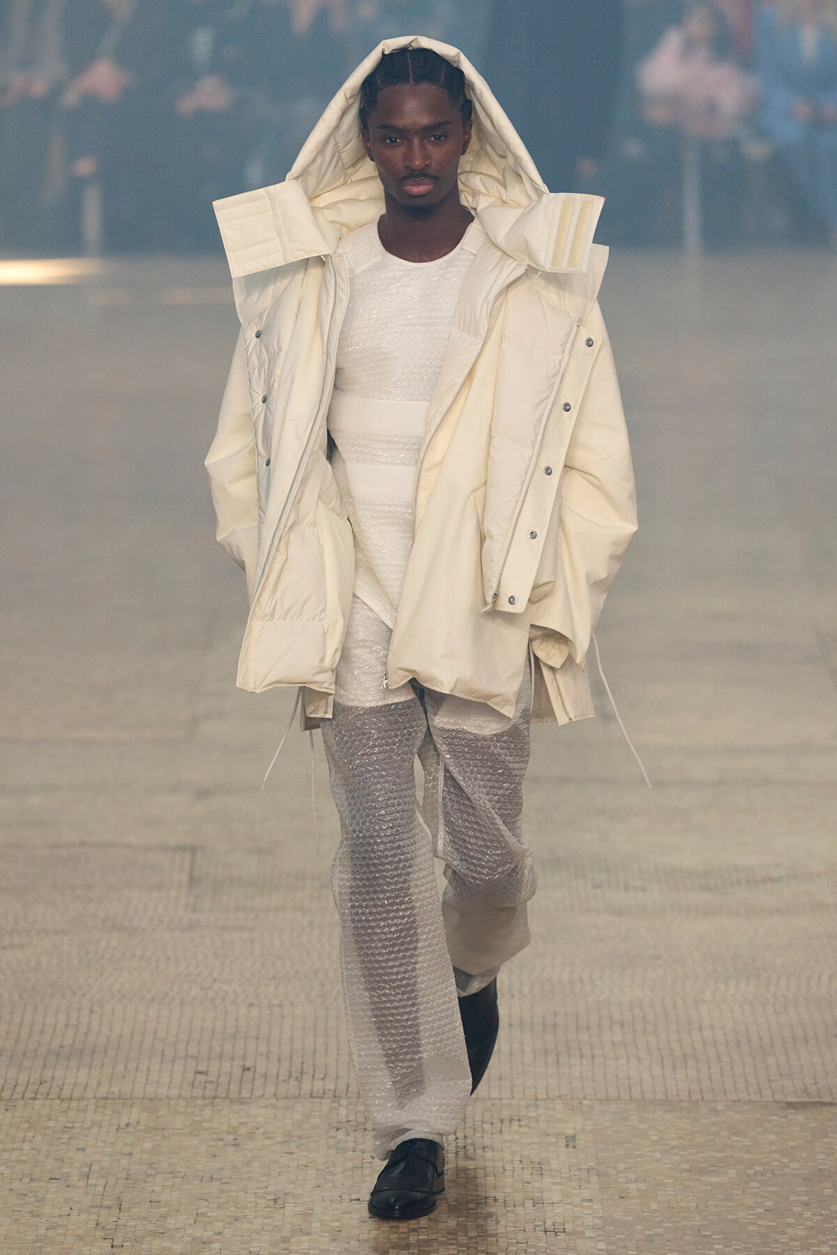 Model on a runway wearing an oversized cream hooded puffer coat over a white textured outfit.