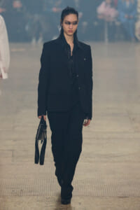 Female model walking a fashion runway in a black pantsuit, carrying a small black handbag, with bold dark eye makeup and a serious expression.