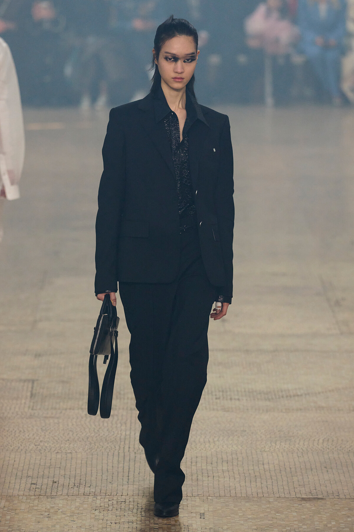 Female model walking a fashion runway in a black pantsuit, carrying a small black handbag, with bold dark eye makeup and a serious expression.