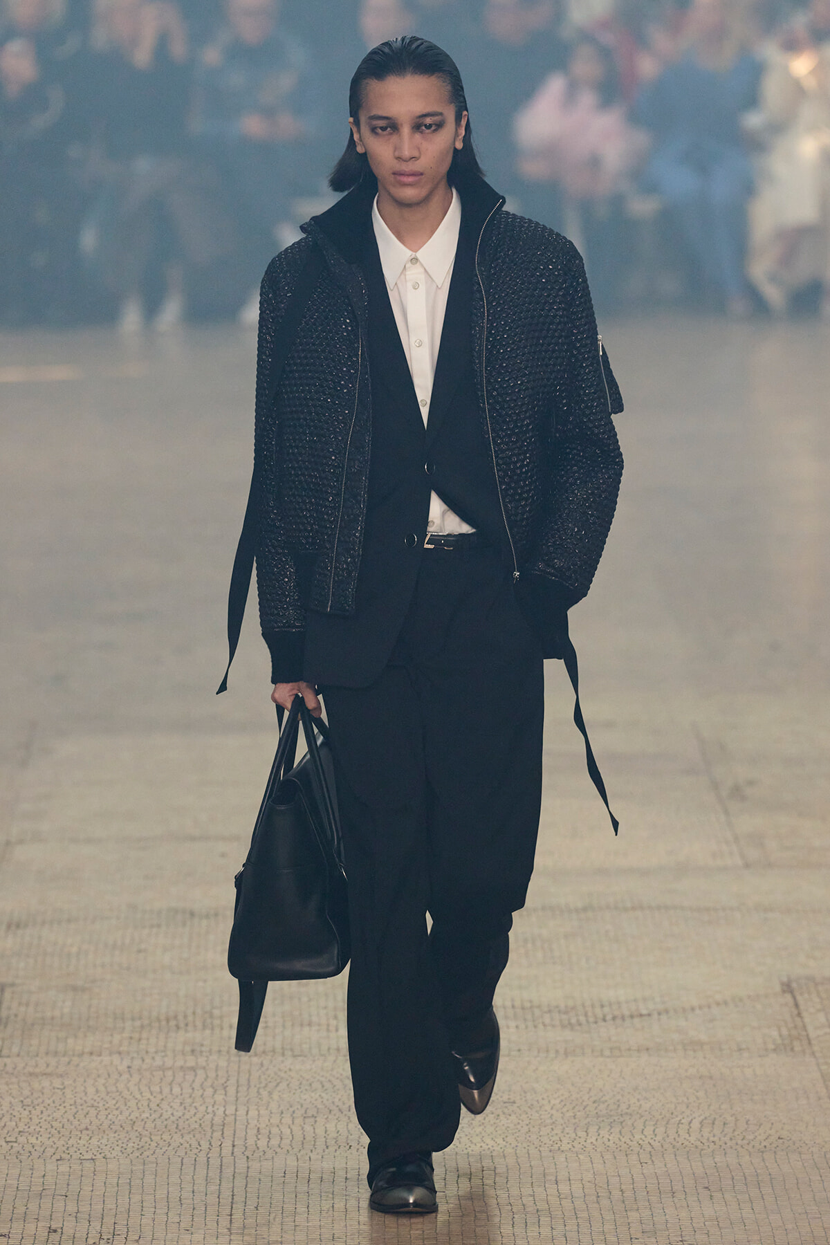 Model walking on a runway in a dark, textured jacket over a white shirt and black suit, carrying a black bag.