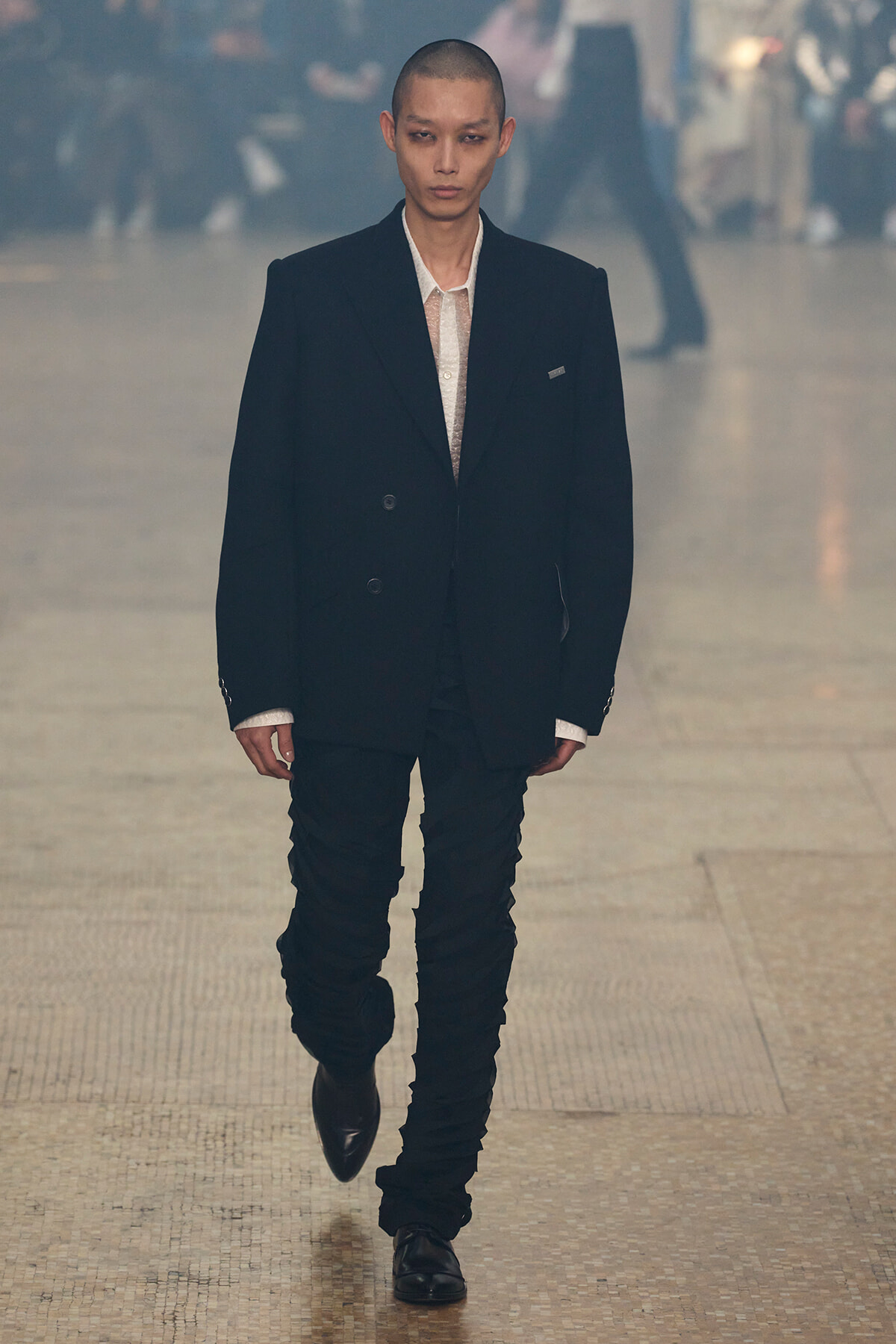 Male model walks a fashion runway wearing a black double-breasted blazer, white shirt, and black pants. Blurred crowd in background.