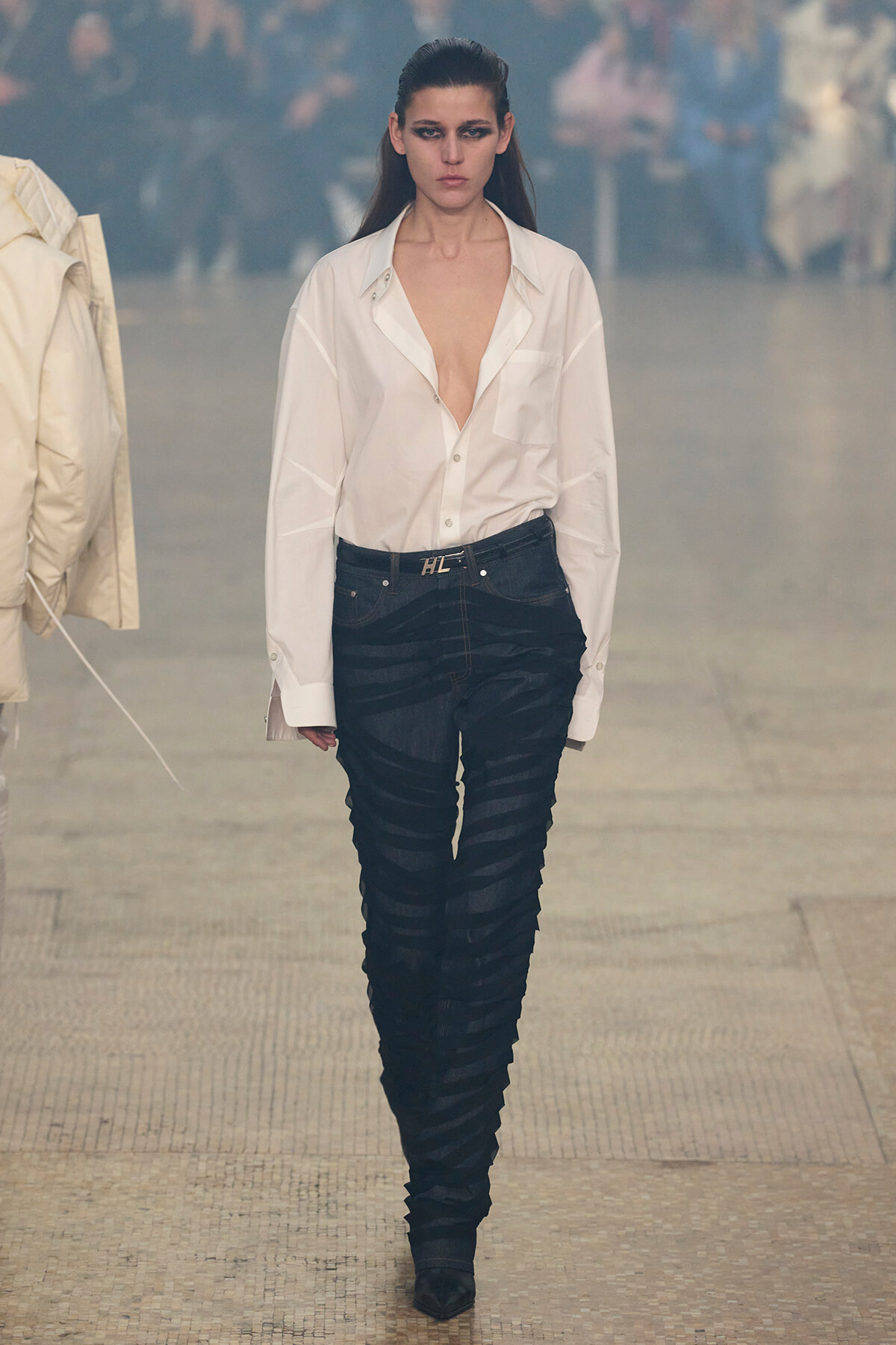 Model walking a runway in a white button-down shirt worn open at the chest and black textured pants with a belt, outdoors with a blurred crowd in the background.