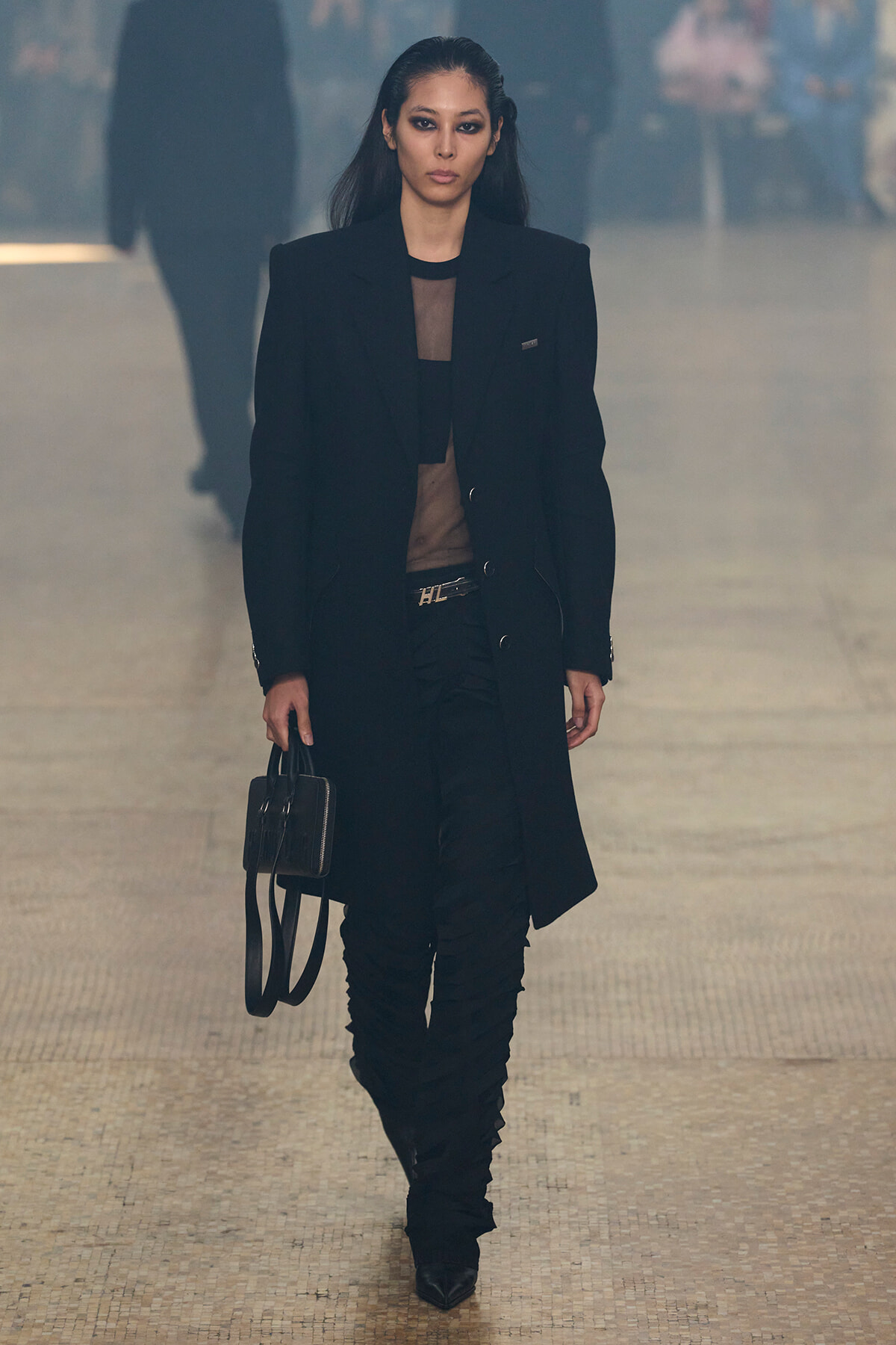 Model walks a fashion runway in a long black coat over a sheer black top, black pants, and heeled boots, carrying a small black handbag