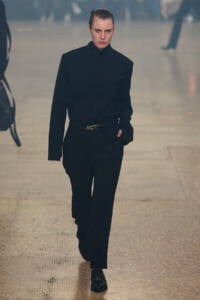 Model walking a runway in an all-black ensemble: high-neck buttoned shirt tucked into tailored trousers with a belt, black laced shoes, serious expression.