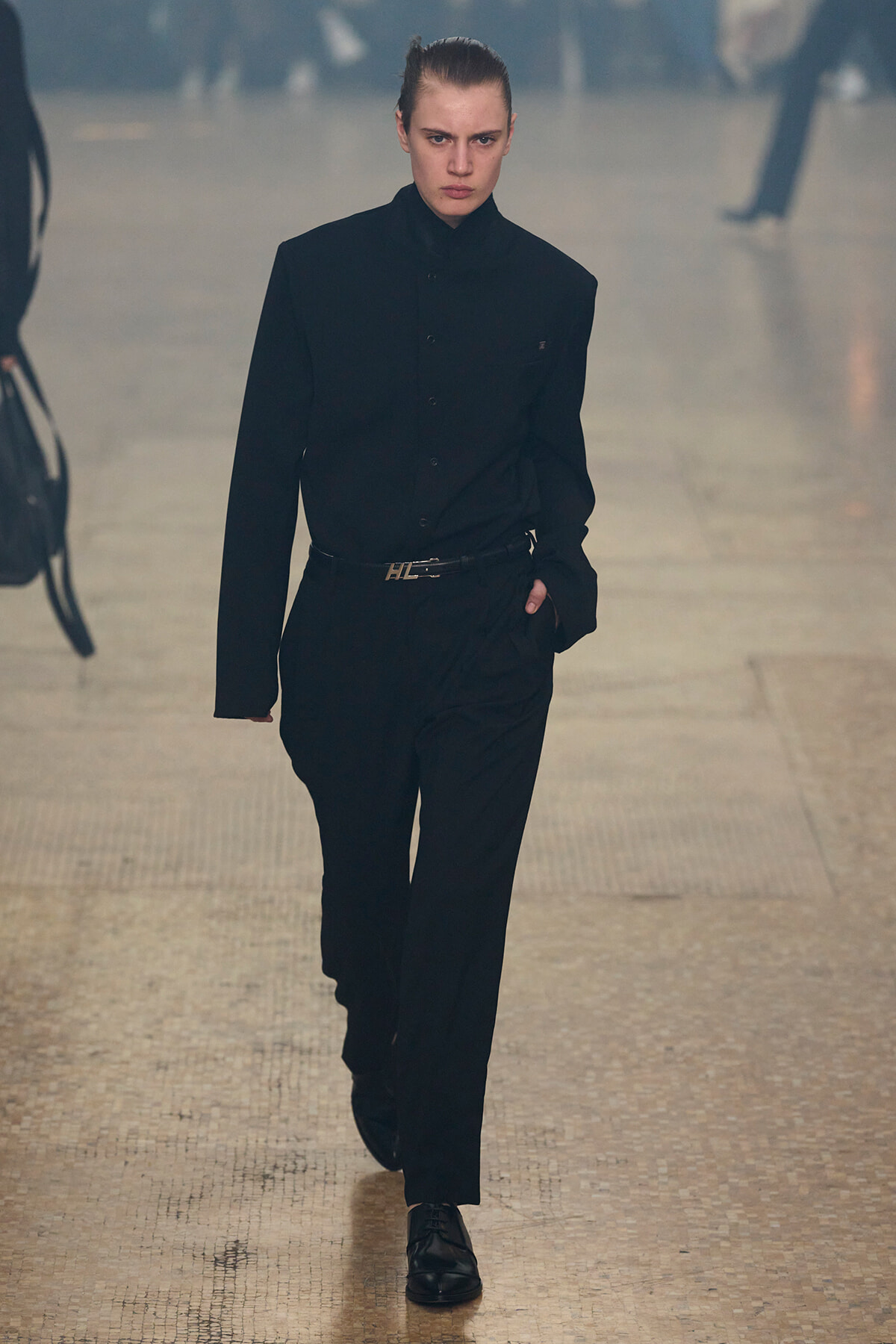 Model walking a runway in an all-black ensemble: high-neck buttoned shirt tucked into tailored trousers with a belt, black laced shoes, serious expression.