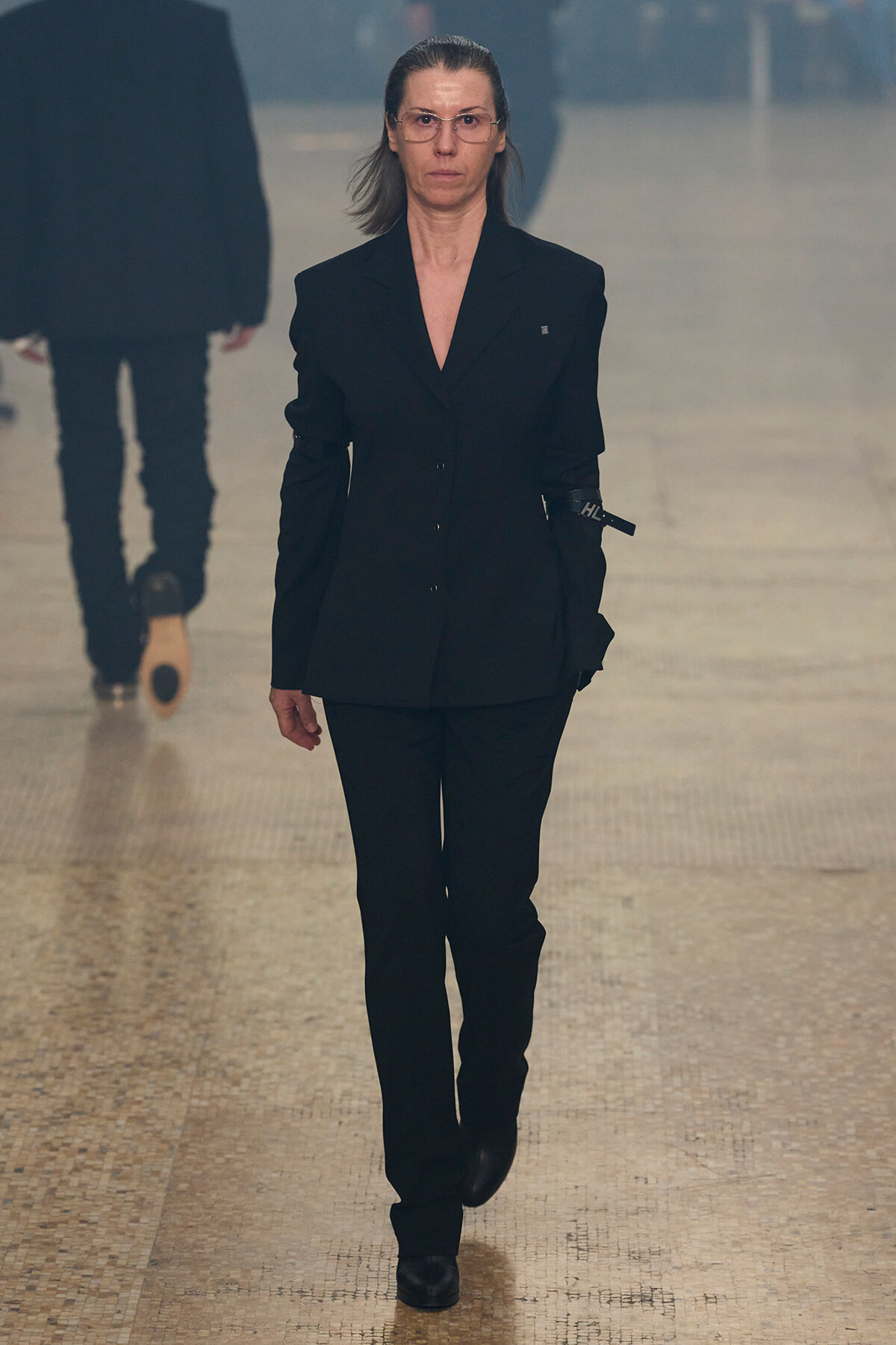 Person walking down a runway, wearing a black tailored suit and boots, with a stern expression.