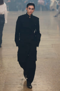 Model walking down a runway in an all-black, structured outfit with a high collar and cargo pockets.