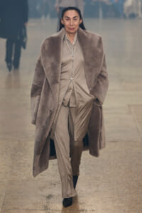 Woman walking toward camera in a large taupe fur coat over a beige satin suit on a wet pavement.