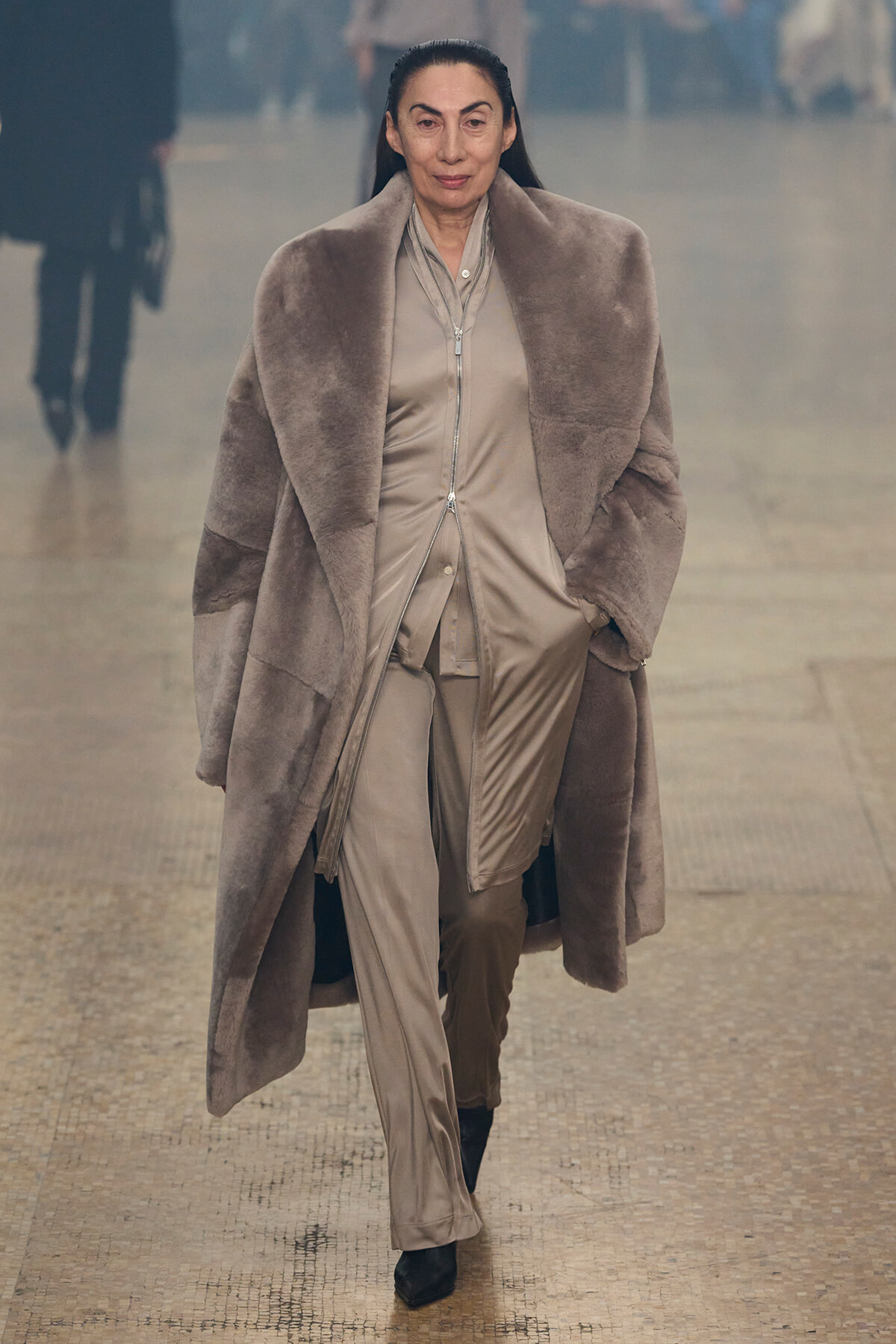 Woman walking toward camera in a large taupe fur coat over a beige satin suit on a wet pavement.