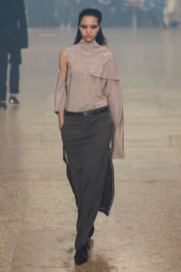 Model walking a runway in a taupe, draped, one-shoulder blouse paired with dark tailored trousers and a slim belt.