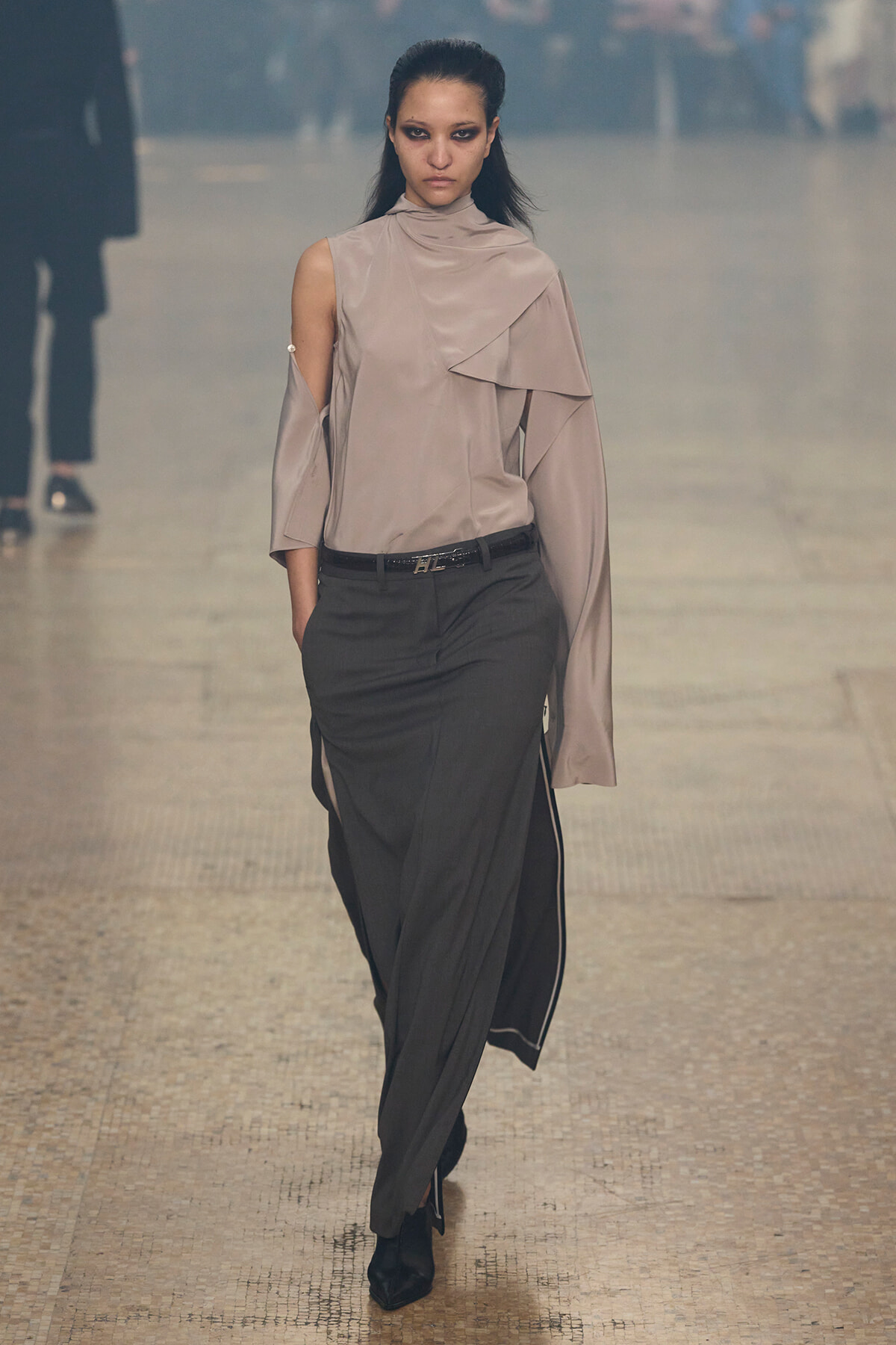 Model walking a runway in a taupe, draped, one-shoulder blouse paired with dark tailored trousers and a slim belt.