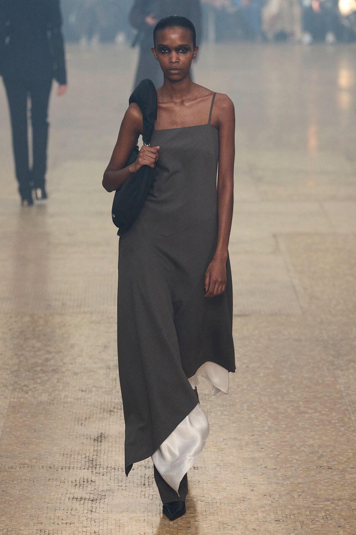 Model walking a runway in a gray asymmetric dress, with a black shoulder bag and black shoes; white fabric peeks from the hem.