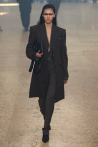 Model walks a runway in a long black blazer with a plunging top, carrying a black clutch/bag under her arm.