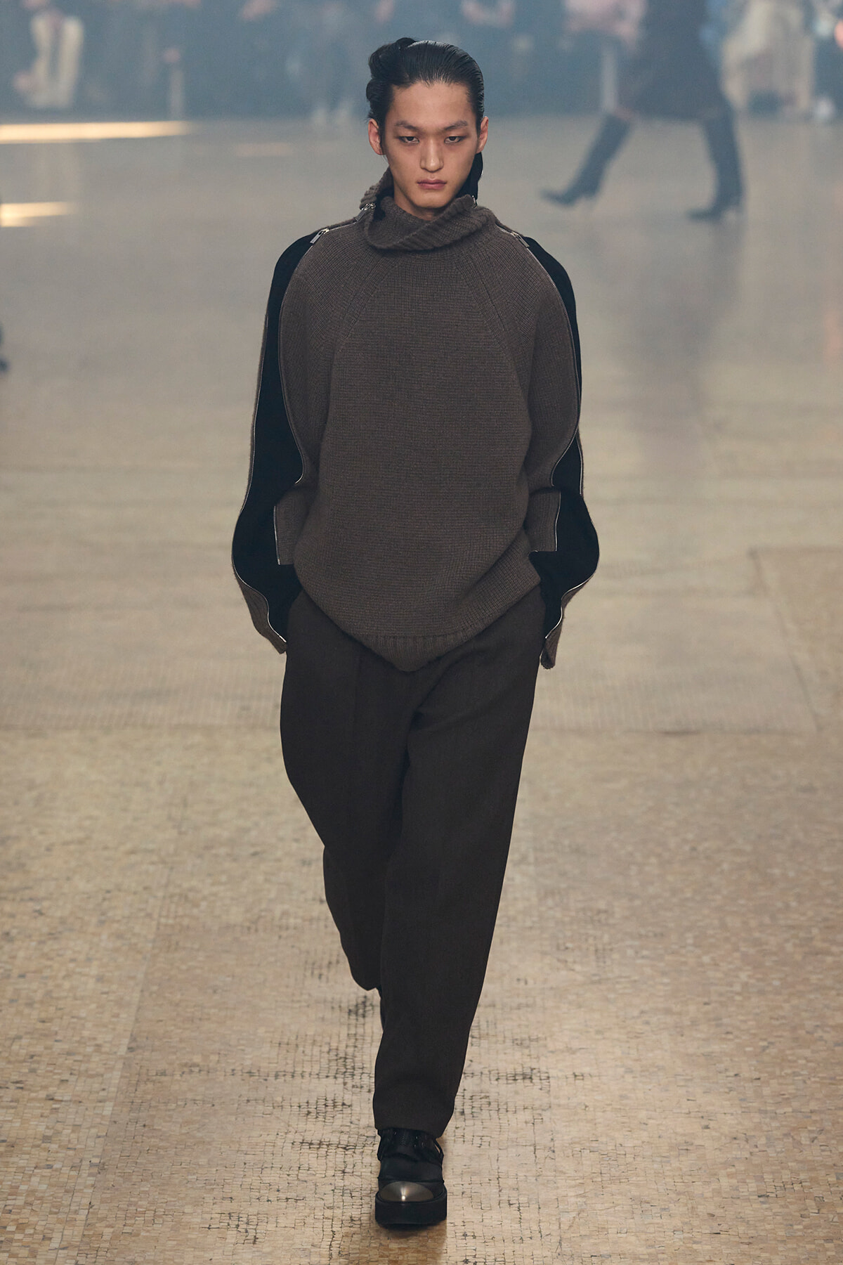 Model walking on a runway in an oversized brown knit turtleneck with black sleeves and brown trousers.