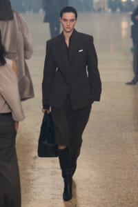 Model walking a fashion runway in a dark oversized blazer suit, knee-high boots, and a black bag.