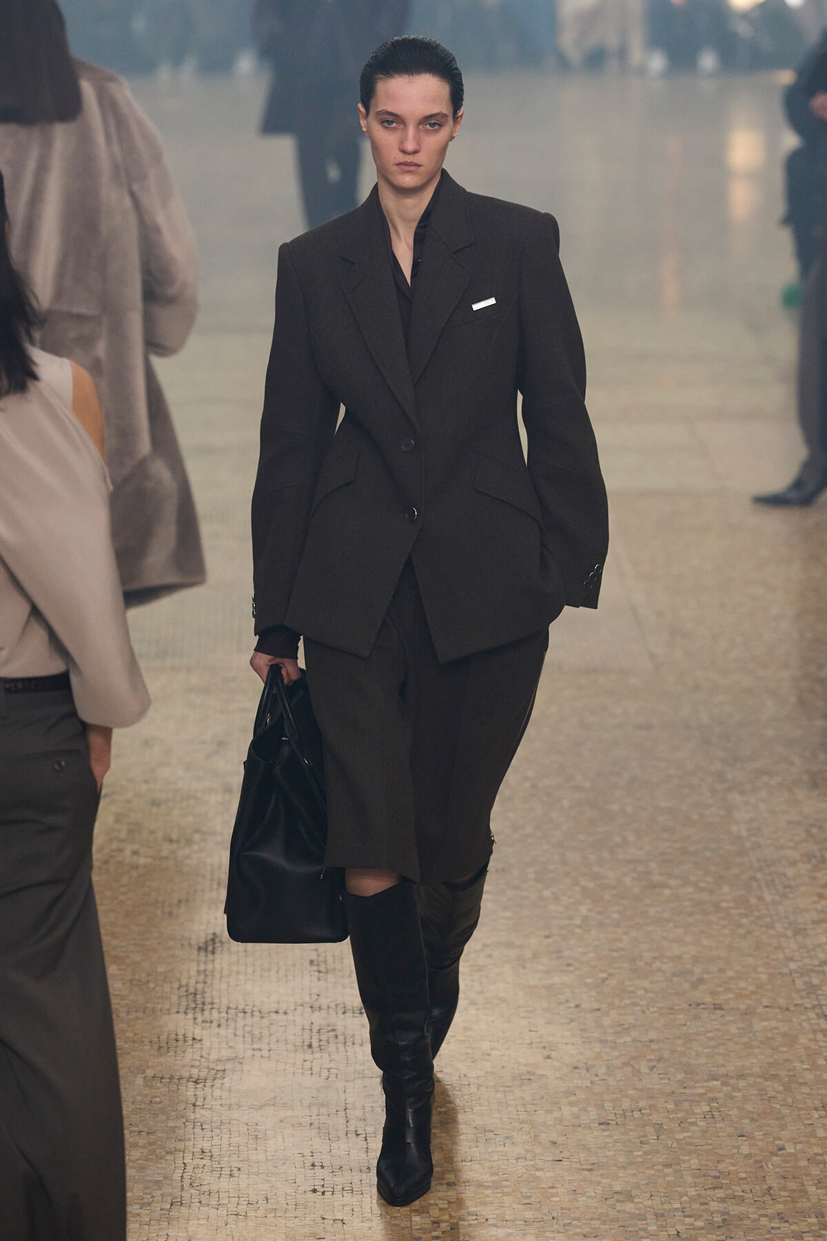 Model walking a fashion runway in a dark oversized blazer suit, knee-high boots, and a black bag.