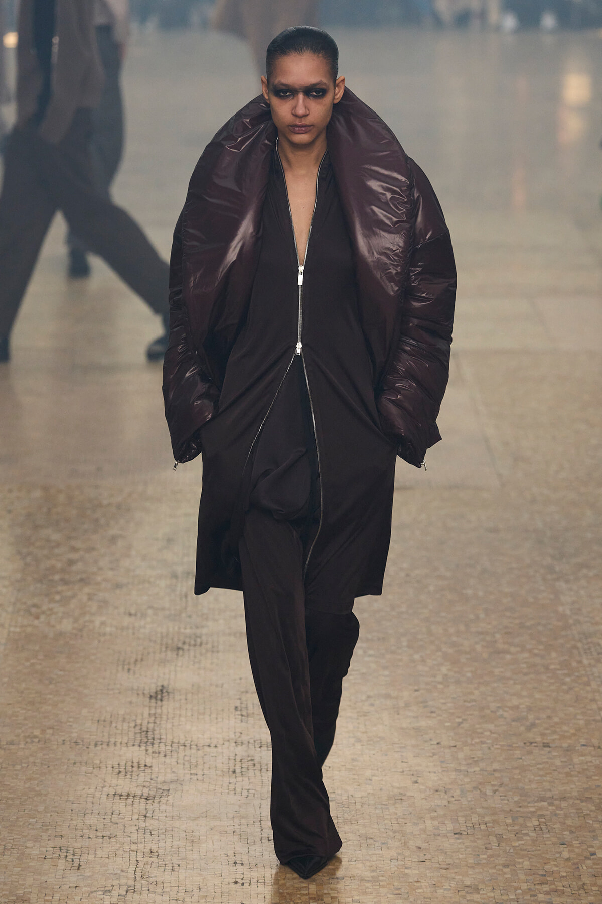 Model wearing an oversized dark brown puffer coat with a high collar, walking on a wet mosaic floor with smoky eye makeup.