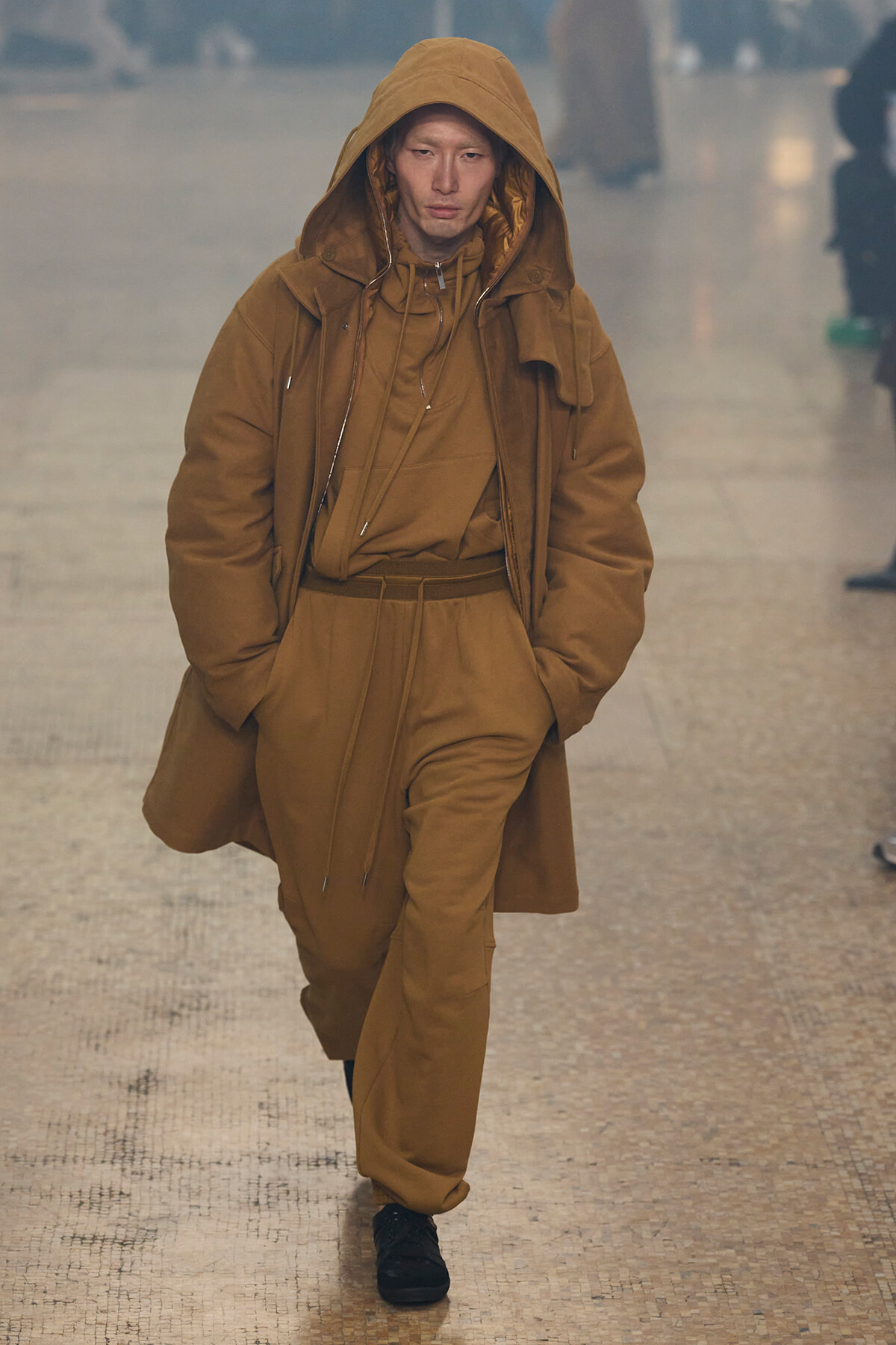 Person in a tan hooded coat and matching pants walking down a tiled indoor walkway in a fashion-forward look.