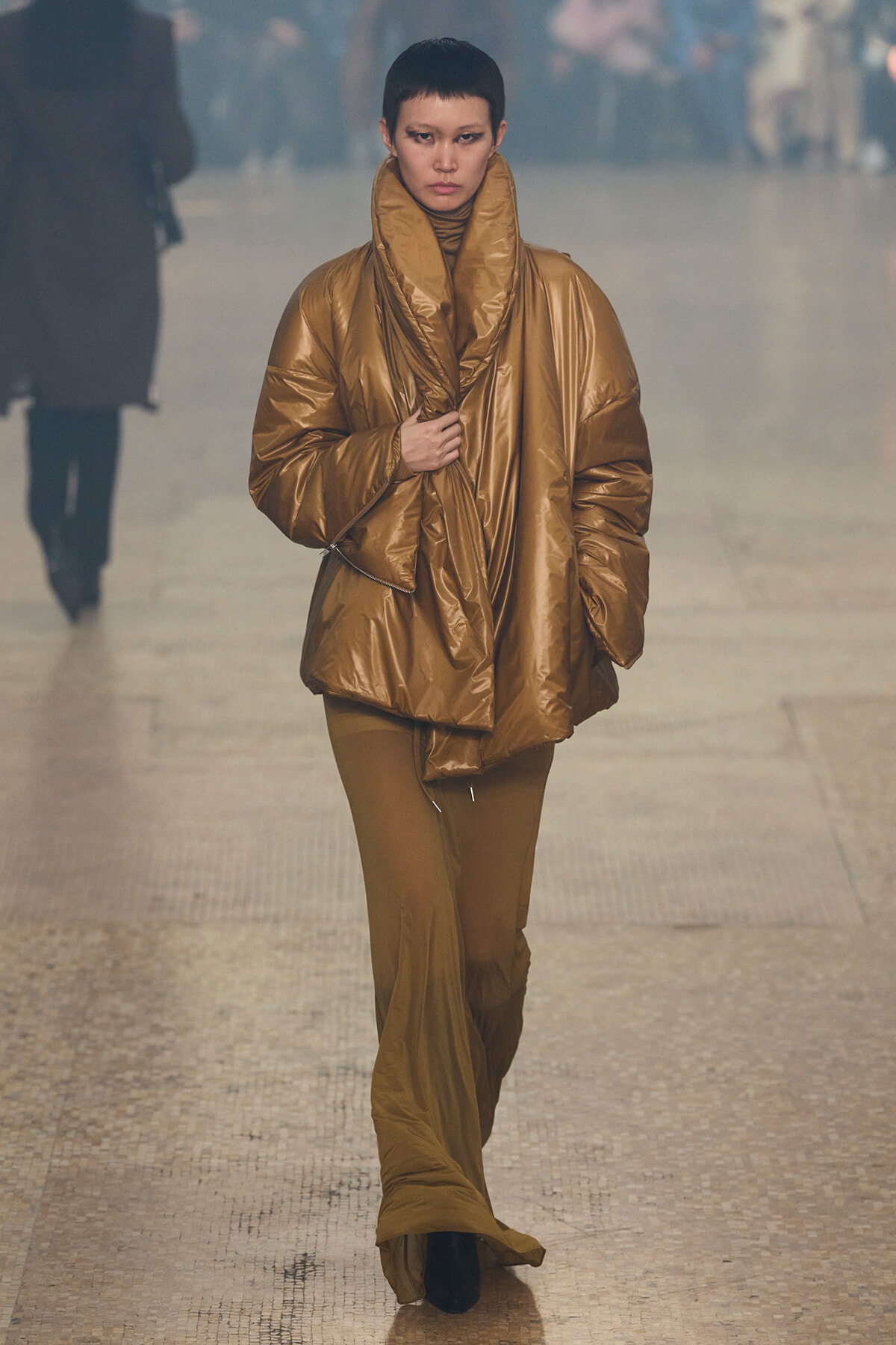 Model walking a fashion runway in a oversized metallic bronze puffer coat with a high, sculpted collar.