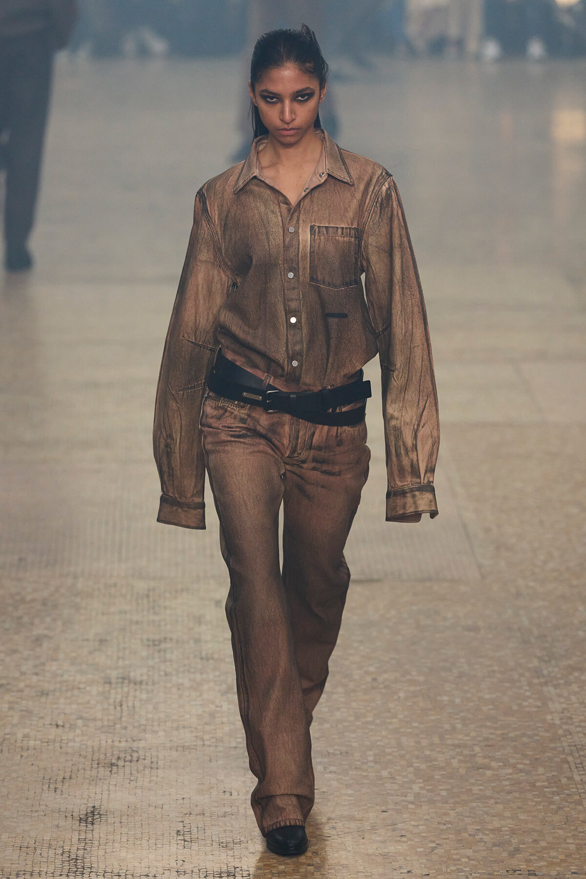Female model walks the runway in a metallic brown shirt and matching pants with a black belt, neutral makeup, and a focused expression.