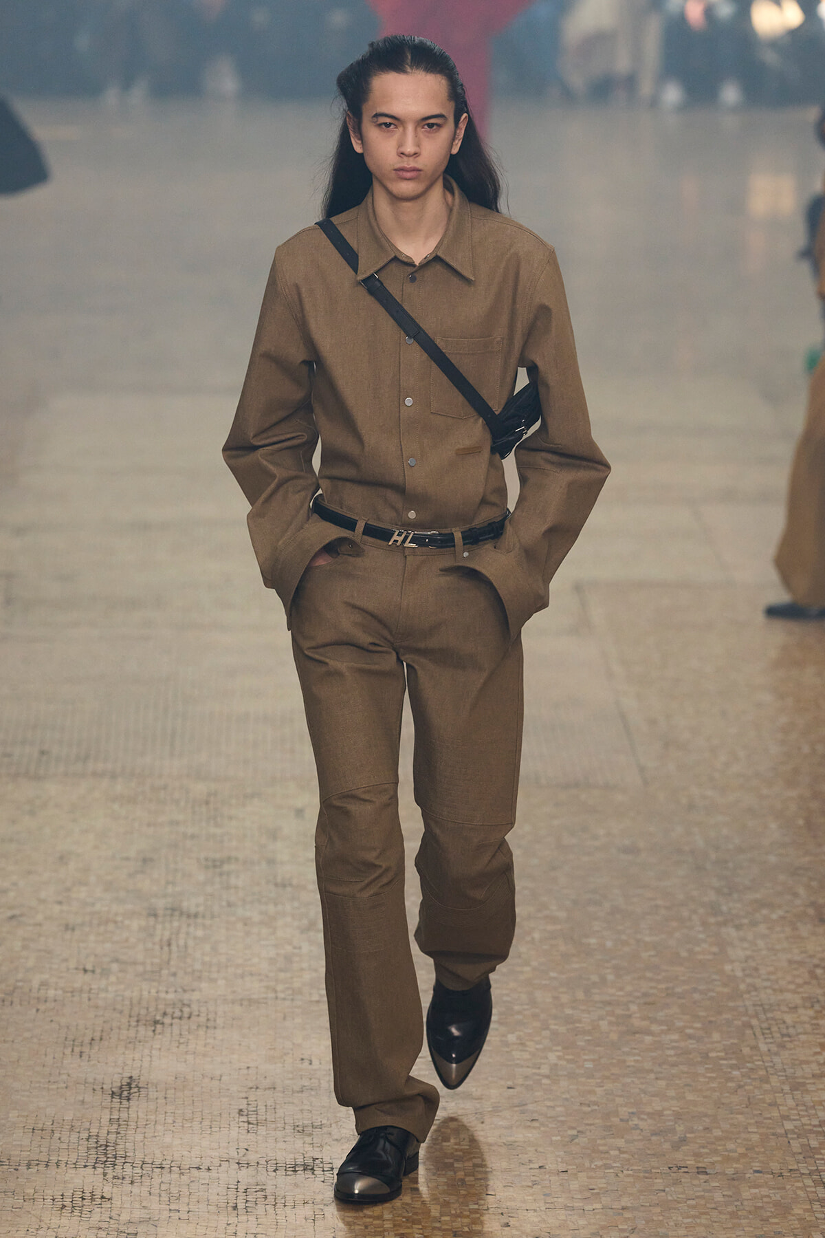 Model walking a runway in a tan matching shirt and pants with a black crossbody bag across the chest