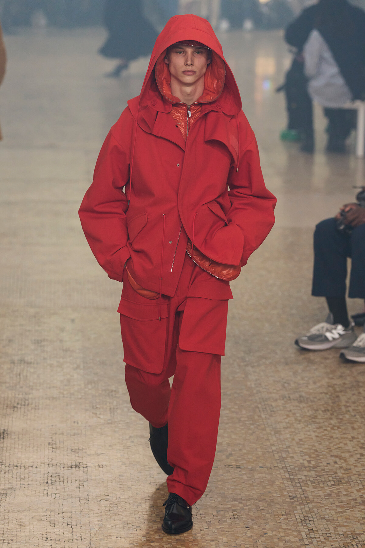 Model on a runway wearing a bright red oversized hooded parka with matching red cargo pants and black shoes.”