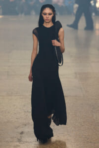 Female model in a long black evening dress with a shoulder bag, walking down a fashion runway.