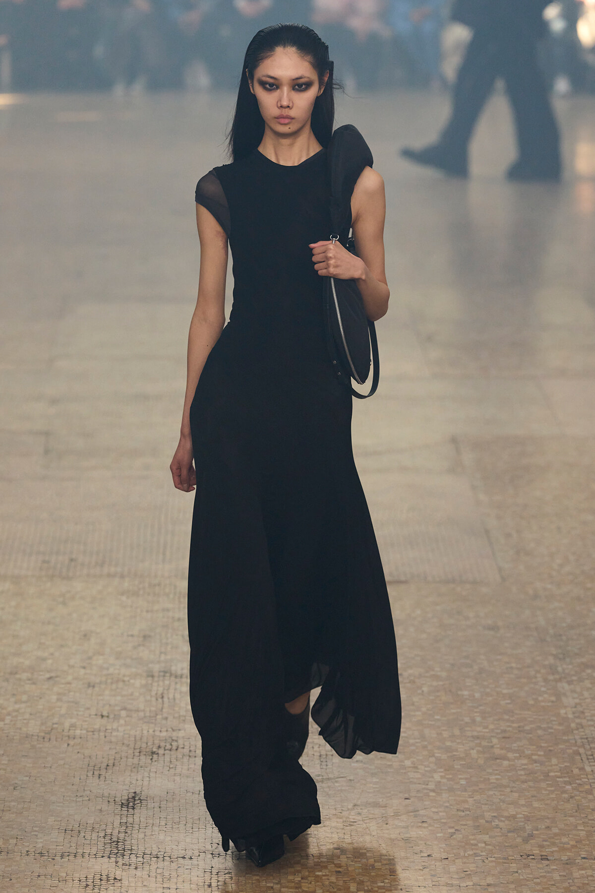 Female model in a long black evening dress with a shoulder bag, walking down a fashion runway.