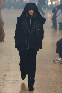 Person wearing a large black hooded parka walking toward the camera in a hazy indoor space.