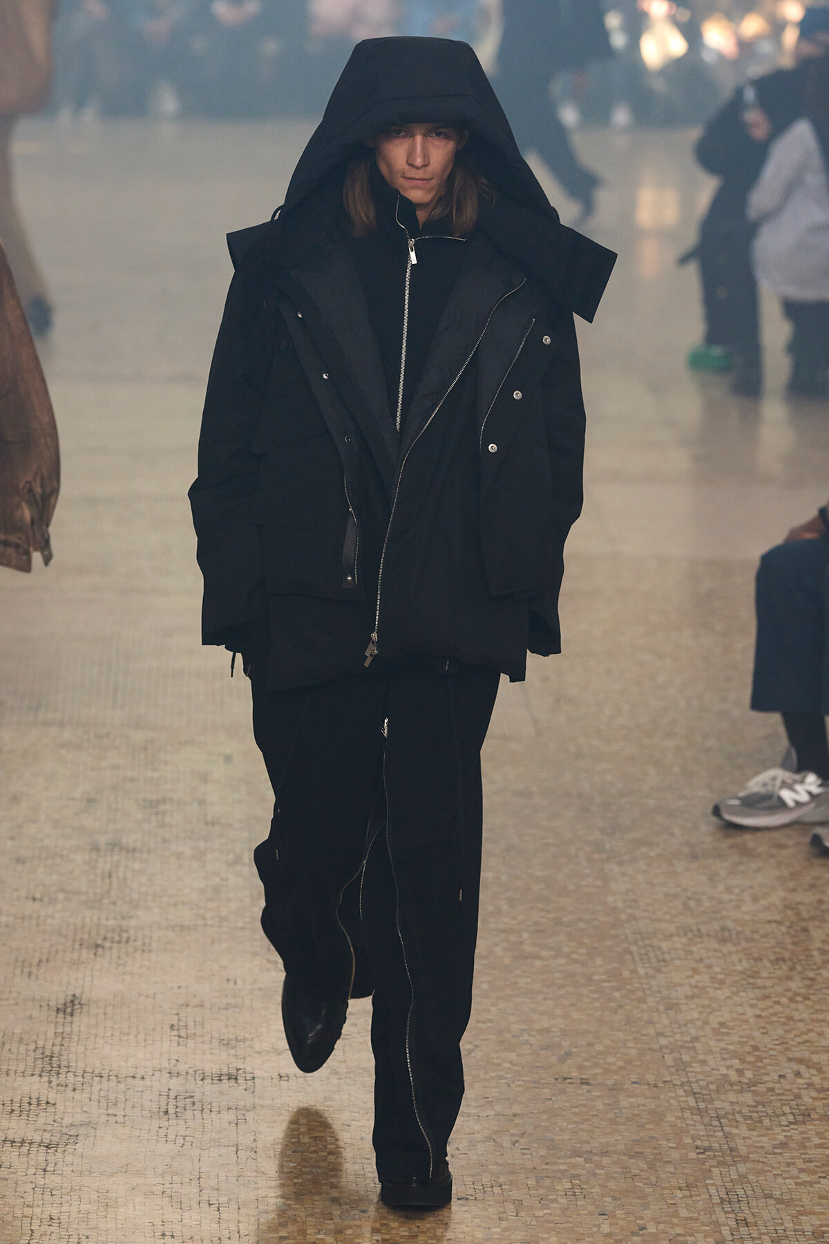 Person wearing a large black hooded parka walking toward the camera in a hazy indoor space.