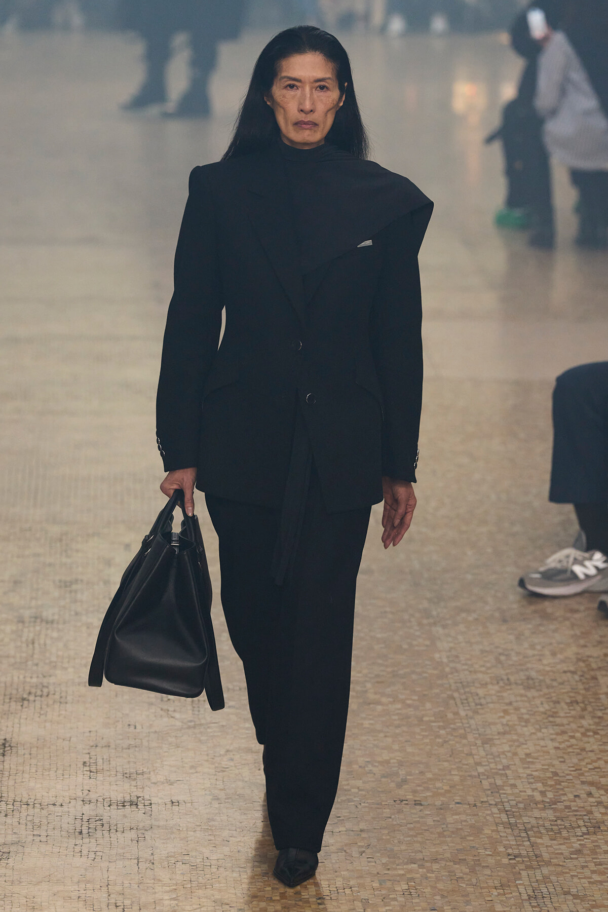 Woman in a black blazer and matching pants walks toward the camera, carrying a large black handbag in a spacious indoor area.