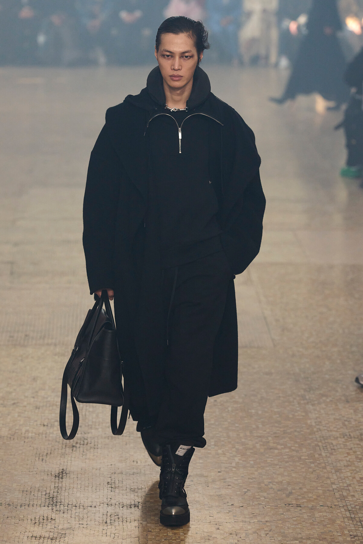 Model walking a runway in an oversized black outfit, carrying a black handbag in hand.