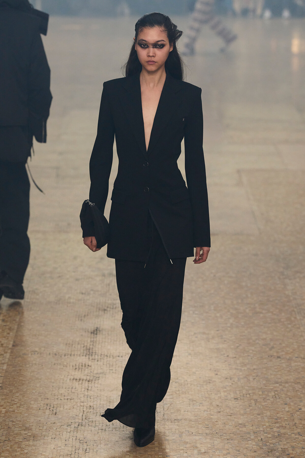Model walks the runway in a black tailored blazer and matching pants, clutch in hand on a beige tiled catwalk.