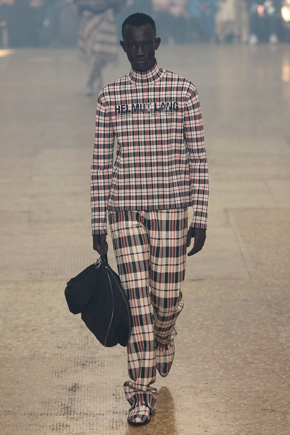 Model wearing a matching black-and-white plaid turtleneck and pants, carrying a black bag on a runway.