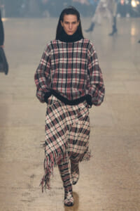 Model walking a runway in a black turtleneck under a large red, black, and white plaid sweater and matching fringed skirt.