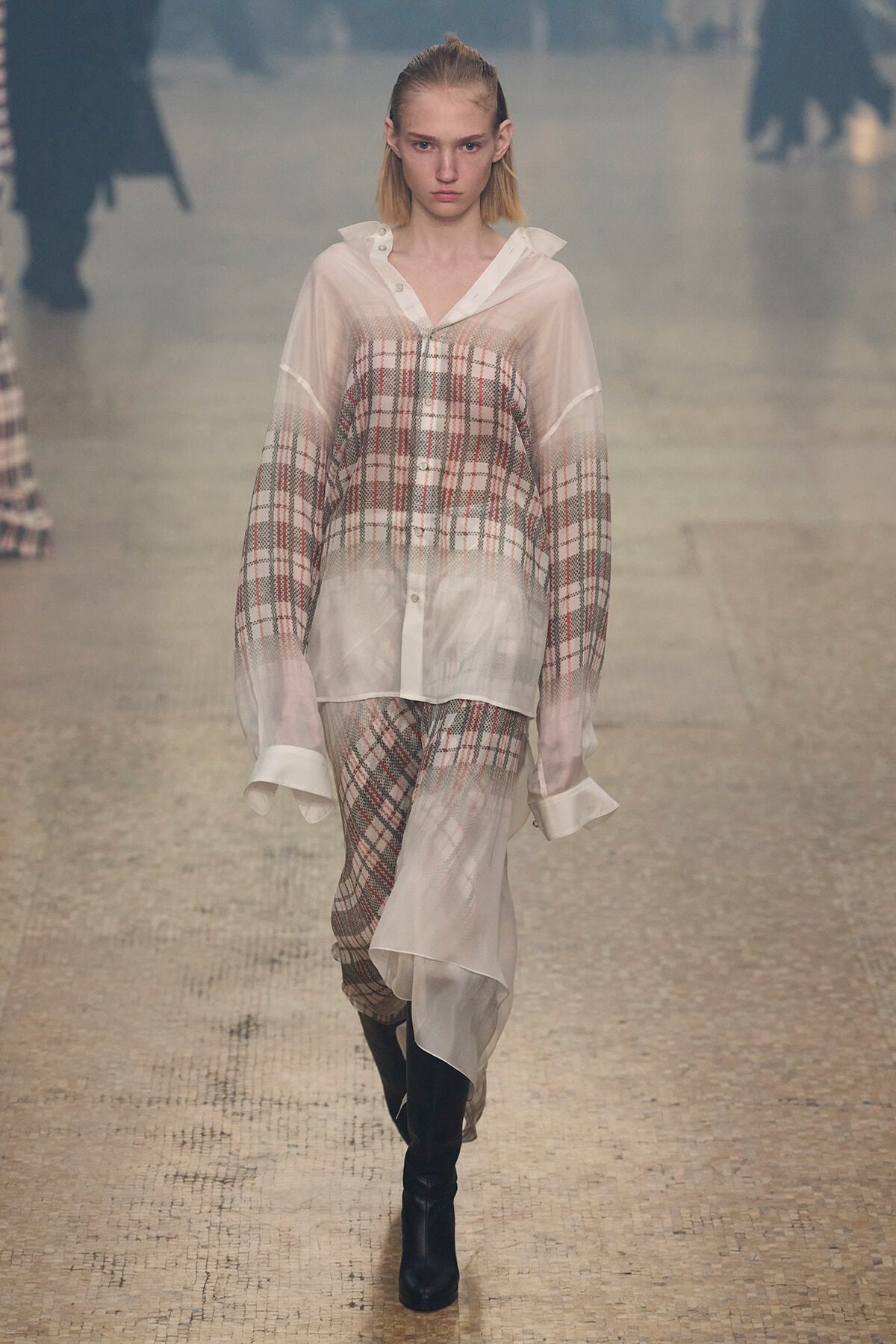 Model walking the runway in a sheer, oversized plaid shirt and matching plaid skirt, paired with black knee-high boots.