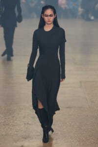 Female runway model in a black long-sleeve draped dress, black boots, and clutch walking down a misty runway.